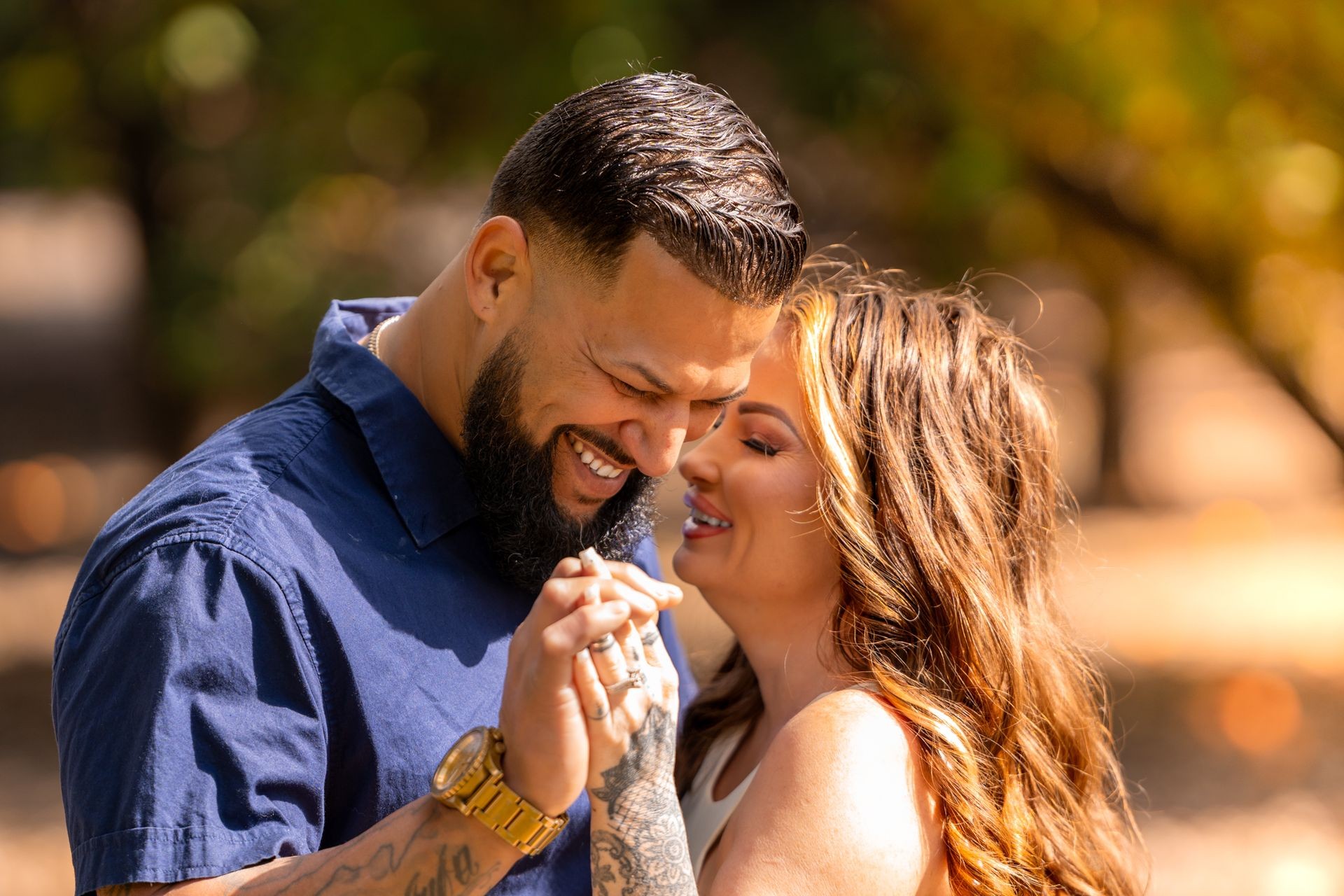 A couple smiling and holding hands while standing close together in an outdoor setting with blurred background.